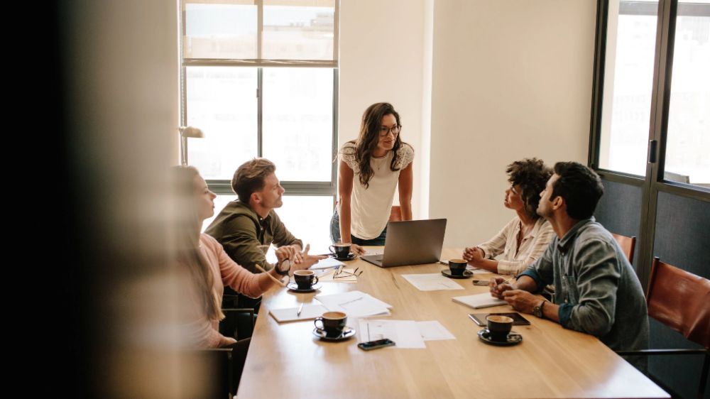 Five professionals having a collaborative meeting around a wooden conference table in a bright office with large windows. A woman wearing a cream-colored top stands while leading the discussion with four seated colleagues, creating an atmosphere of active engagement and teamwork.