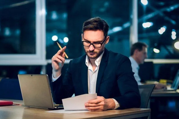 A focused businessman in glasses reviews paperwork at his desk with a laptop while holding a pencil in a dimly lit modern office. 