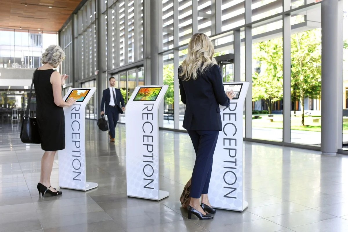Two women use digital reception kiosks in a modern glass-walled lobby while a businessman walks through the bright hallway. 